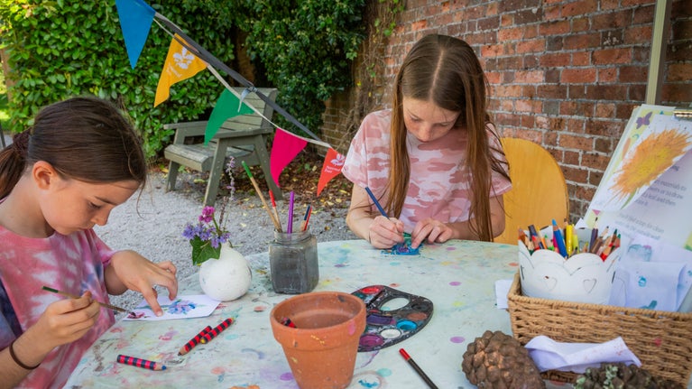 Two girls sat at a crafting table with paints, crayons and paper busy working on their crafts